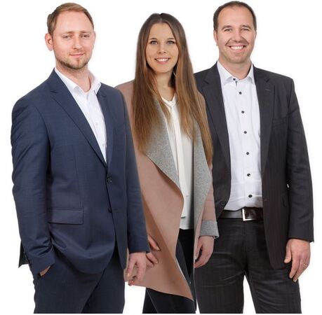 Drei Geschäftsleute in Businesskleidung stehen lächelnd vor weißem Hintergrund.